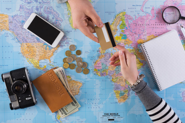 Reisekreditkarte wird überreicht über einer Landkarte auf der sich ein Fotoapparat, ein Smartphone, ein Block und ein Reisepass mit einigen Scheinen Bargeld befindet